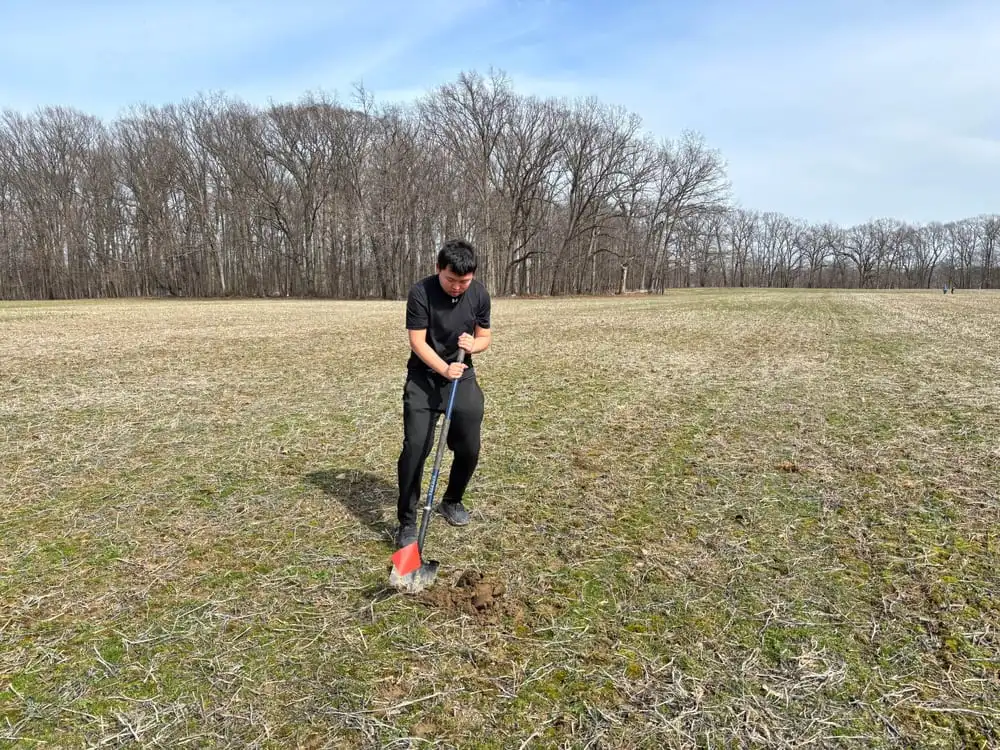 A student breaking ground with a spade in a bare field in front of a winter treeline