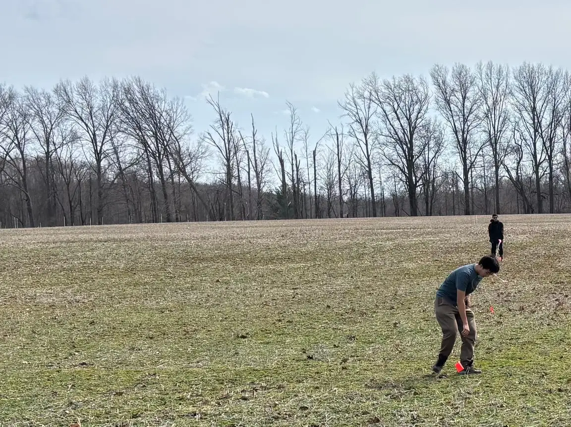 Two students marking planting spots with flags in an open field at the edge of the woods